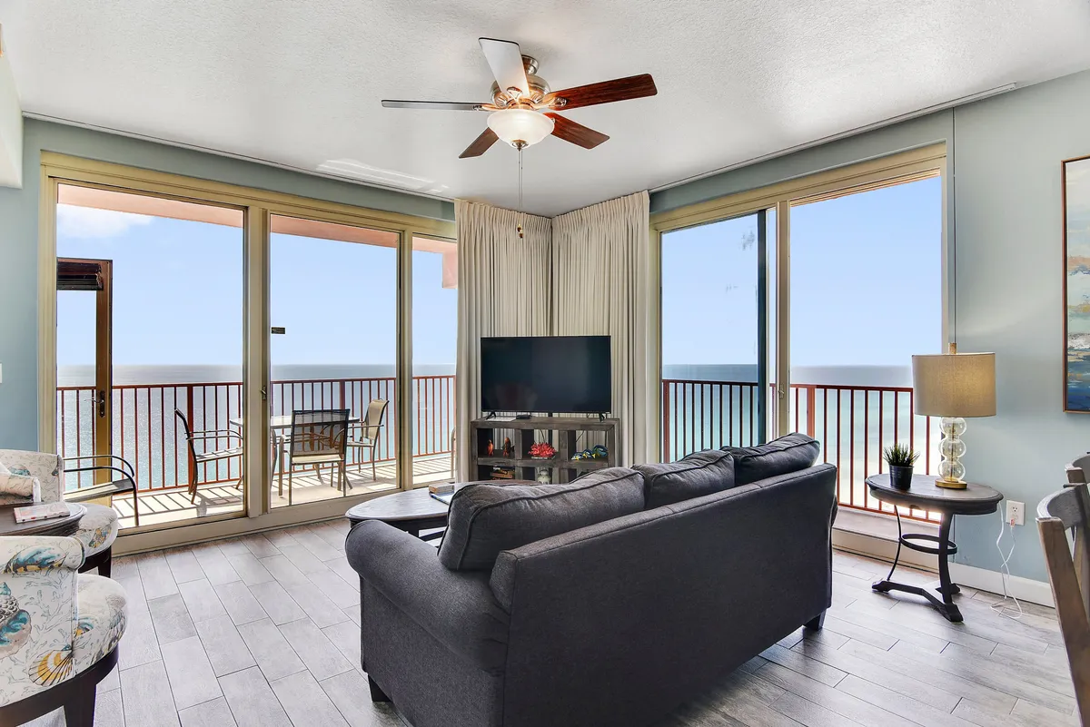 A spacious living area with a dark gray sofa facing a large flat-screen TV, ceiling fan overhead, and panoramic floor-to-ceiling windows that open onto a wraparound balcony with outdoor seating. The view from the windows and balcony reveals stunning Gulf vistas and abundant natural light fills the room, enhancing the contemporary decor.