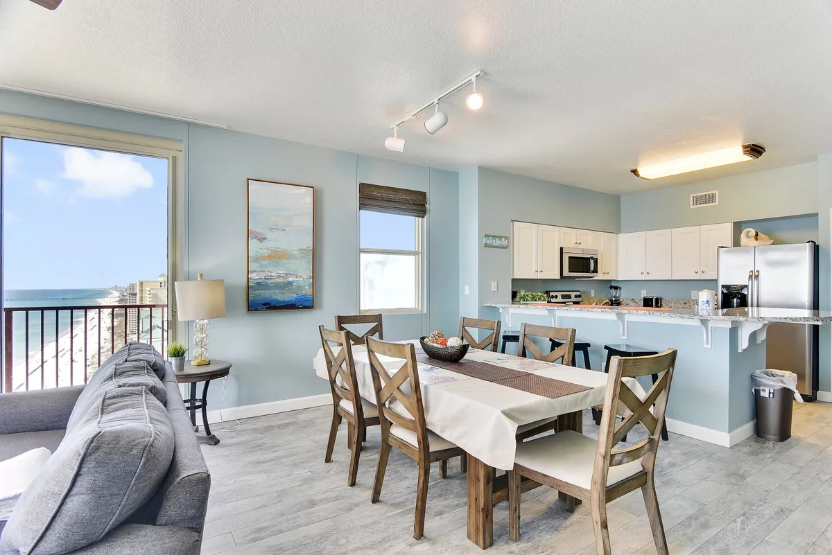 A spacious dining and kitchen area with a dining table that seats six, granite countertops, modern appliances, and a large window and balcony door offering panoramic beach views. The neutral-toned sofa adjacent to the dining area creates a seamless flow between gathering, dining, and relaxing.
