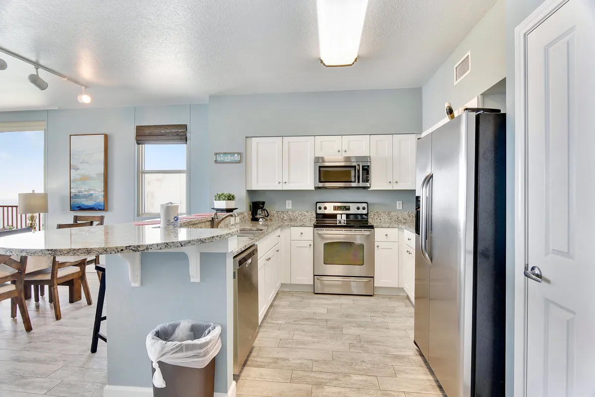 A bright, airy kitchen featuring granite countertops, stainless steel appliances, a breakfast bar with stools, and an adjacent dining area with large windows showcasing an ocean view. The space is open and modern, with ample natural light that emphasizes the kitchen’s inviting atmosphere and its connection to the dining and living areas.