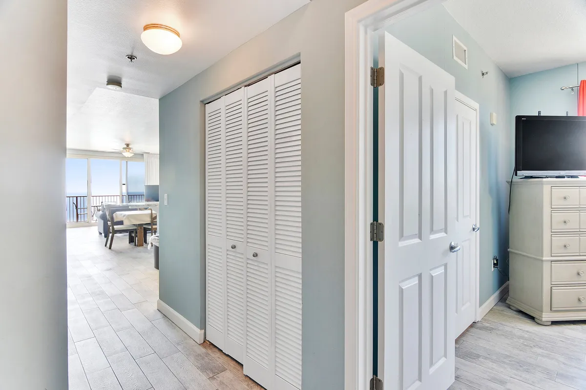 A calm, inviting hallway leads toward the living area, with a glimpse of the balcony and ocean beyond. White louvered closet doors and soft blue walls create a welcoming entryway. Part of a bedroom and dresser with TV are visible, suggesting a cohesive flow from entry to living spaces.