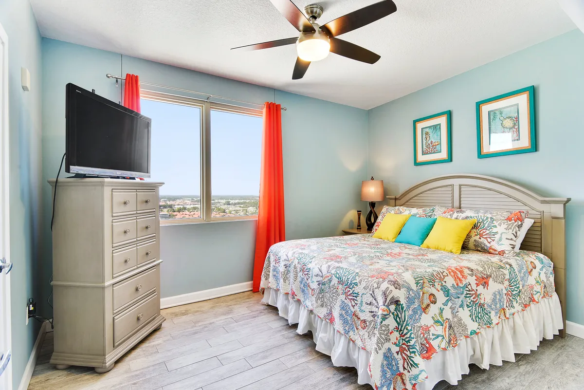 A cheerful and relaxed bedroom with a king-sized bed adorned in colorful coral-themed bedding and bright coral curtains. The room includes a tall dresser with a flat screen TV and two framed ocean-themed artworks above the bed, creating a cozy and inviting atmosphere.