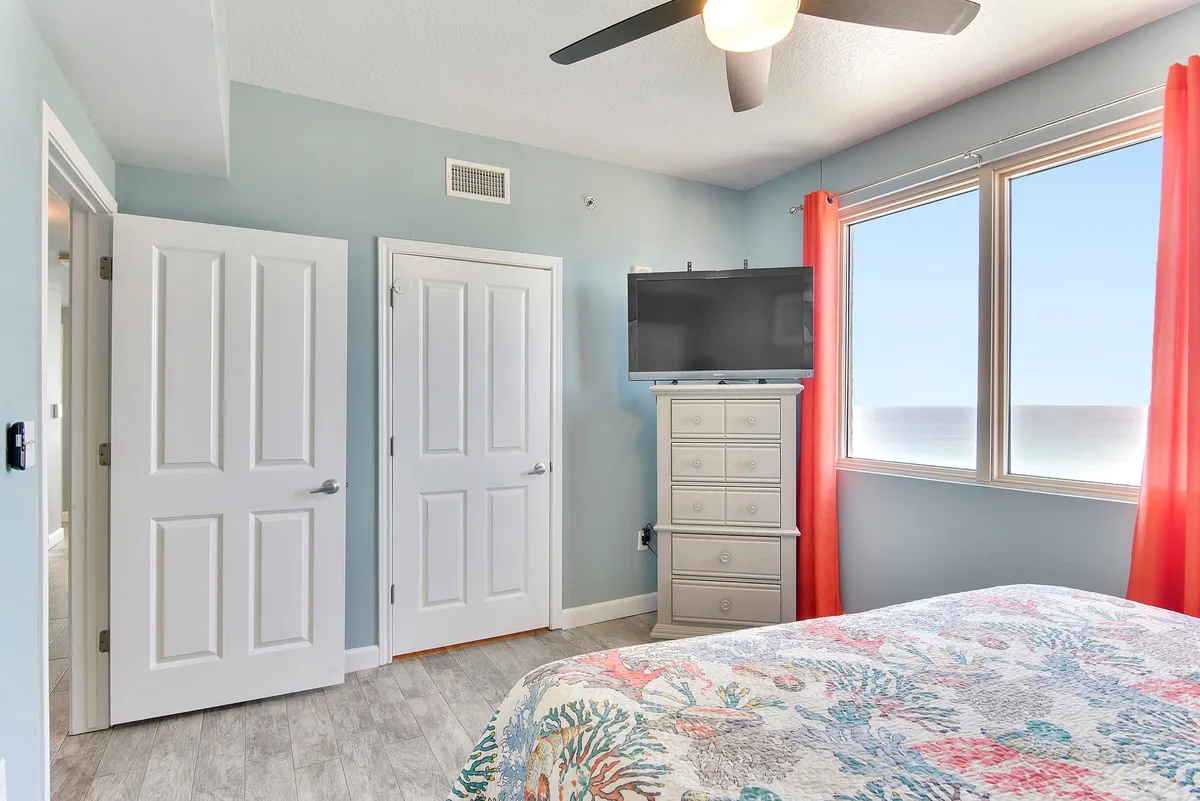 A serene master bedroom featuring a king size bed with colorful coral and ocean-themed bedding. The room boasts a large window showcasing a breathtaking panoramic gulf view, a flat screen TV mounted above a white dresser, and a ceiling fan with a light fixture. The bright, airy atmosphere is enhanced by natural light pouring in from the window.