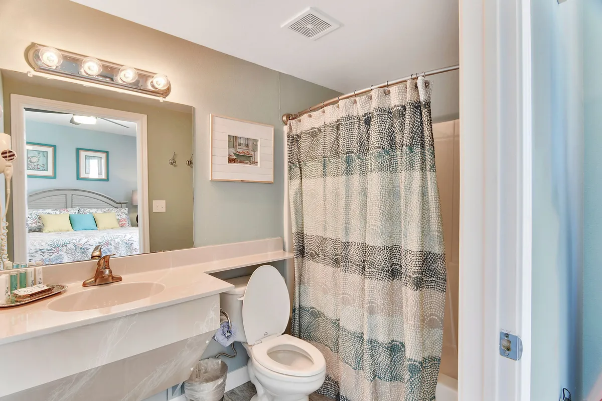 A fresh, inviting bathroom featuring a bathtub/shower combo with a patterned curtain, a long white vanity with integrated sink and bronze faucet, a large mirror reflecting the adjacent bedroom, and a toilet. The space is practical and bright, ideal for post-beach routines.