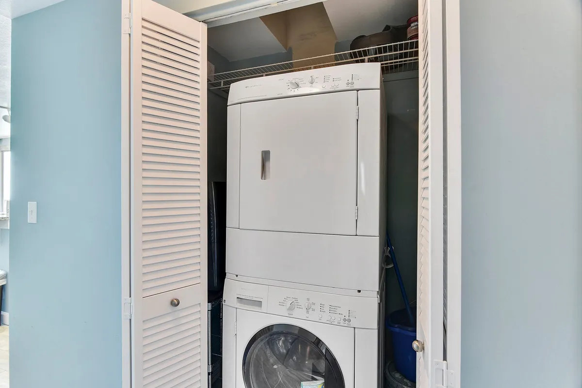 A practical laundry area with a stacked washer and dryer, white louvered closet doors, and clean blue painted walls. A metal wire shelf holds cleaning supplies, highlighting convenience and functionality for guests managing beachwear and towels.