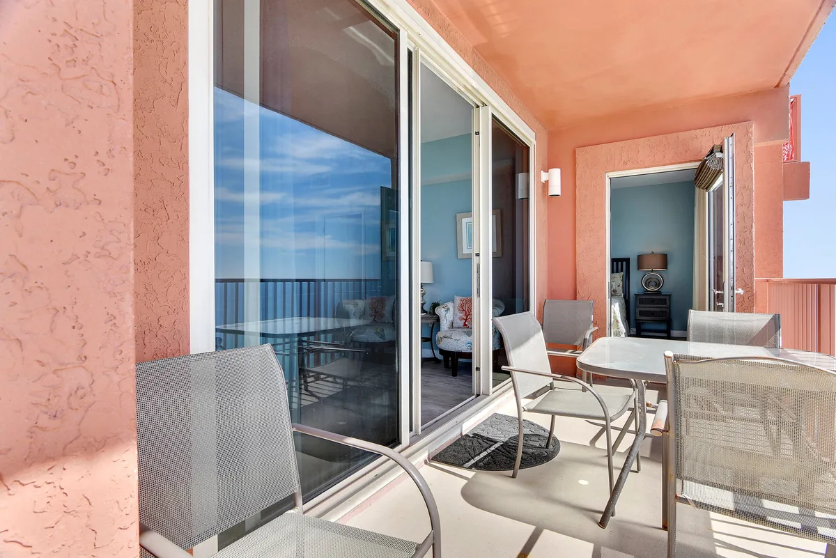 A view from inside the condo looking out through large sliding glass doors onto a wraparound balcony with seating. The balcony offers sweeping views of the ocean, and the reflection in the glass hints at the bedroom's proximity. The scene connects indoor living with private outdoor enjoyment.