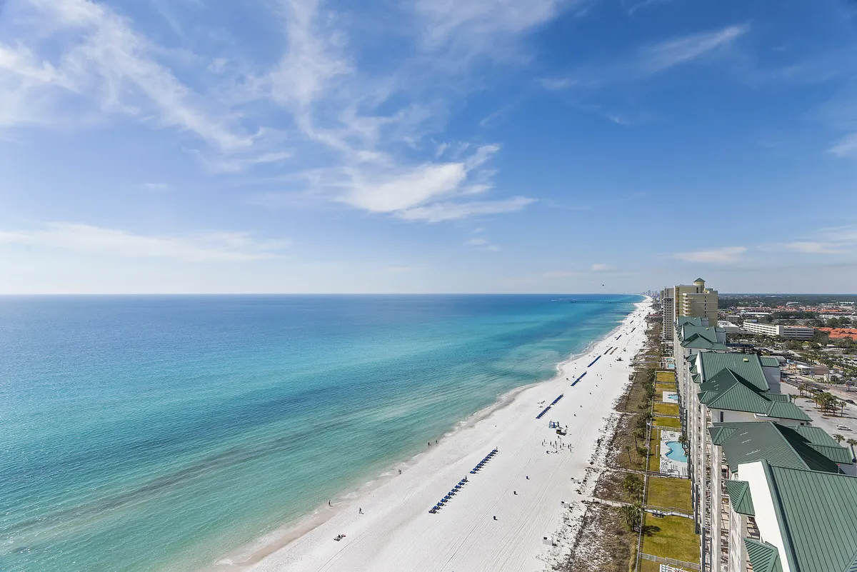A panoramic exterior view of pristine white sandy beaches stretching along turquoise gulf waters, with a line of beach umbrellas and a series of resort buildings in the background. The inviting shoreline is dotted with guests enjoying sunbathing and beach activities.