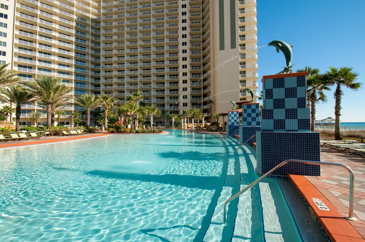 A sprawling lagoon-style swimming pool with playful dolphin water features, set against the backdrop of a tall resort building and lush palm trees. Sun loungers line the pool deck, inviting relaxation and social gatherings. The scene radiates a tranquil vacation vibe, blending water recreation with resort luxury.