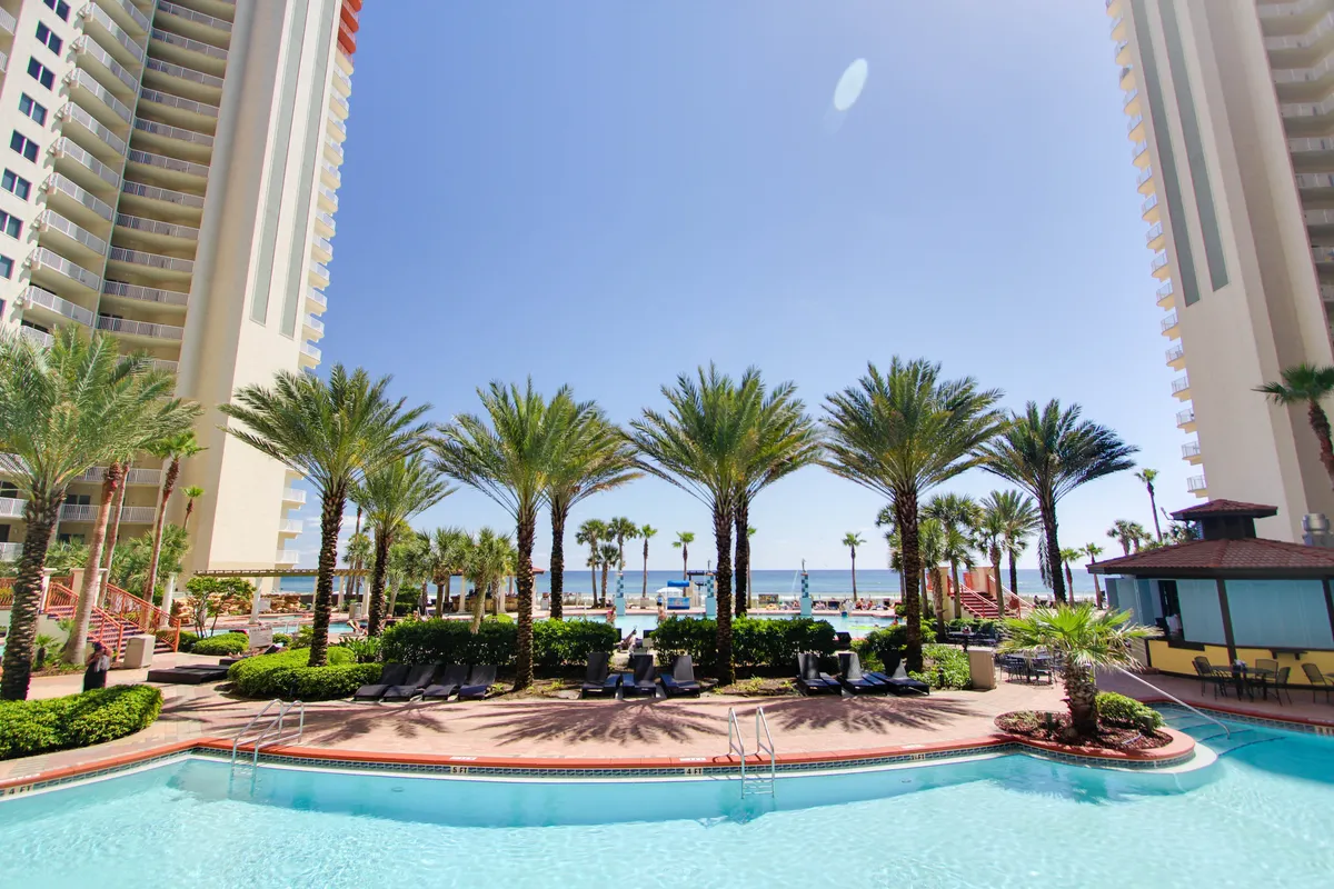 A large lagoon-style swimming pool is surrounded by tall palm trees, with two high-rise resort buildings framing the scene. The sparkling pool water reflects the sky, and the ocean and sandy beach are visible beyond the pool area. The space is lively yet serene, evoking a tropical resort atmosphere.