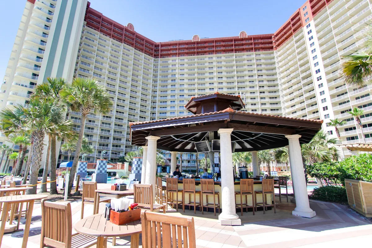 The photo showcases a central tiki bar gazebo with seating, surrounded by wooden tables and chairs, with a large resort building and tropical landscaping in the background. The scene is casual yet upscale, conveying a relaxed social environment perfect for evening cocktails or casual gatherings.