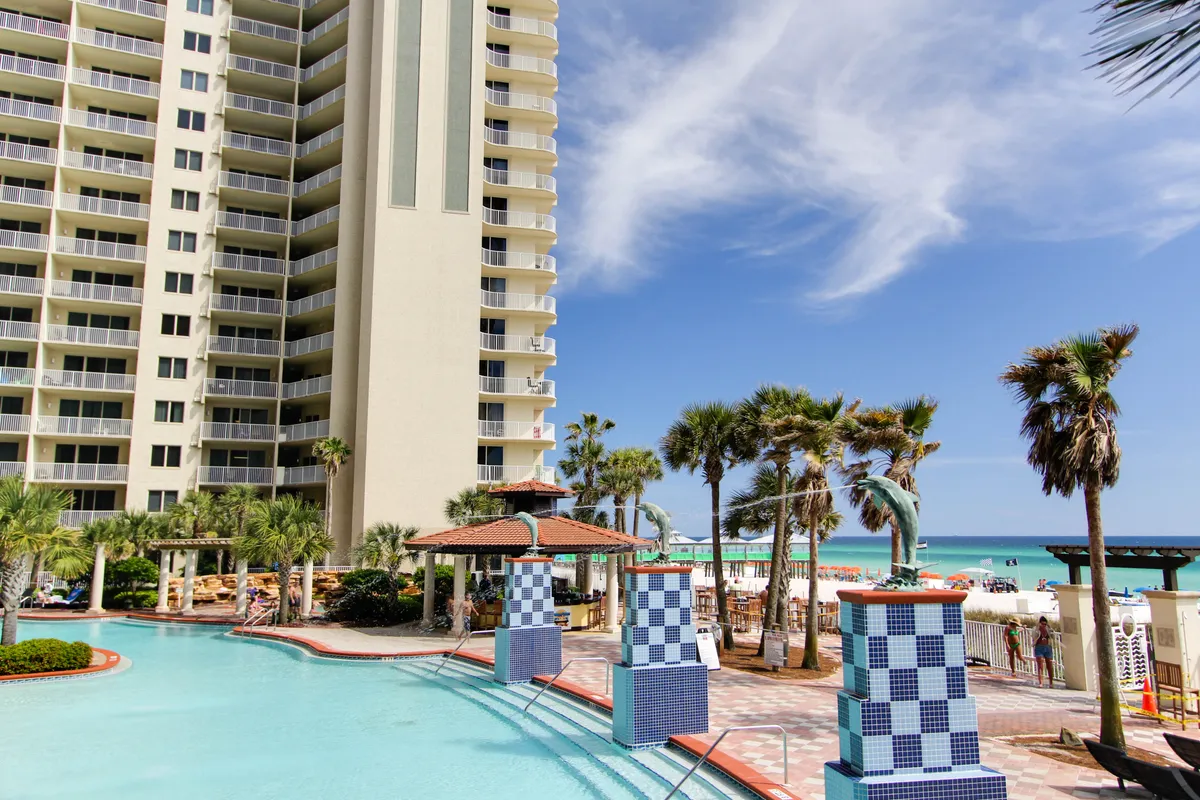 This exterior shot captures a sprawling lagoon-style swimming pool, a tiki bar with a vibrant red roof, and a multi-story resort building with balconies. Palm trees and the ocean are visible in the background, emphasizing the tropical, beachfront atmosphere.