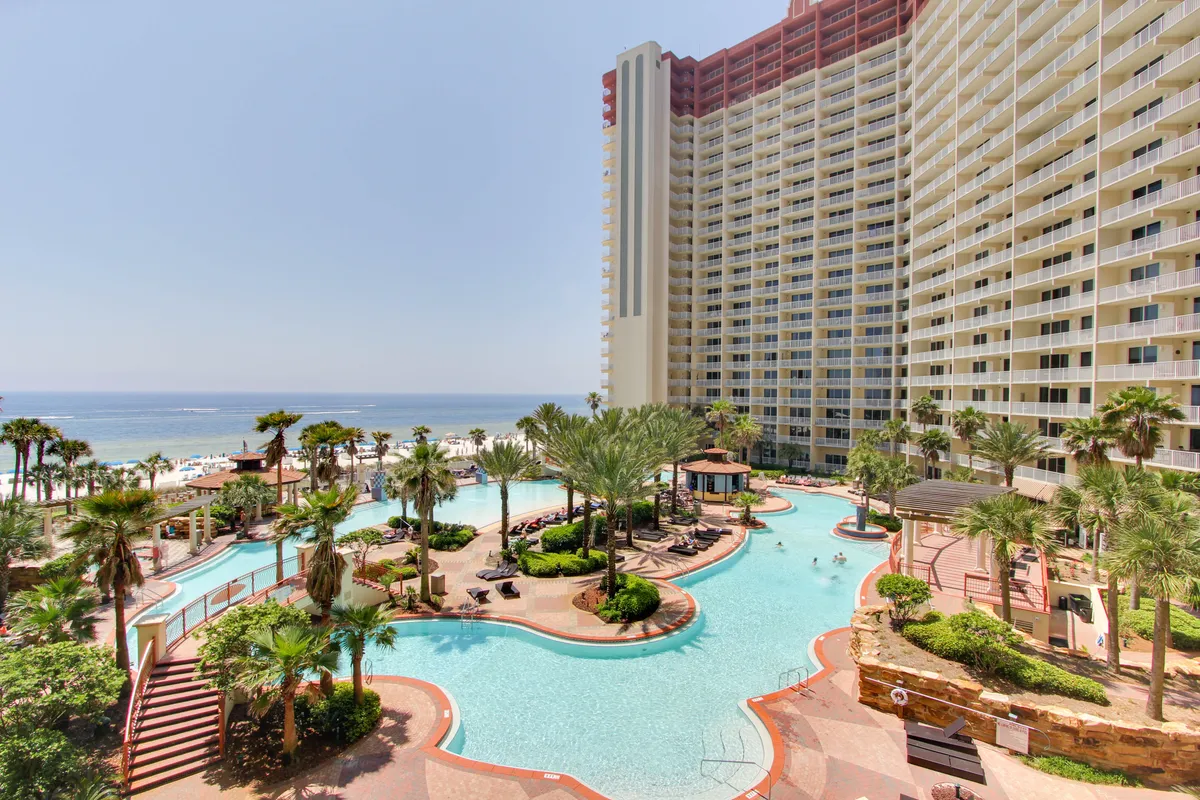 A large lagoon style swimming pool is surrounded by lush palm trees and landscaped greenery, with a tall, multi-story resort building featuring numerous balconies in the background. The pool area includes water fountains and is adjacent to a sandy shoreline, signaling private beach access.