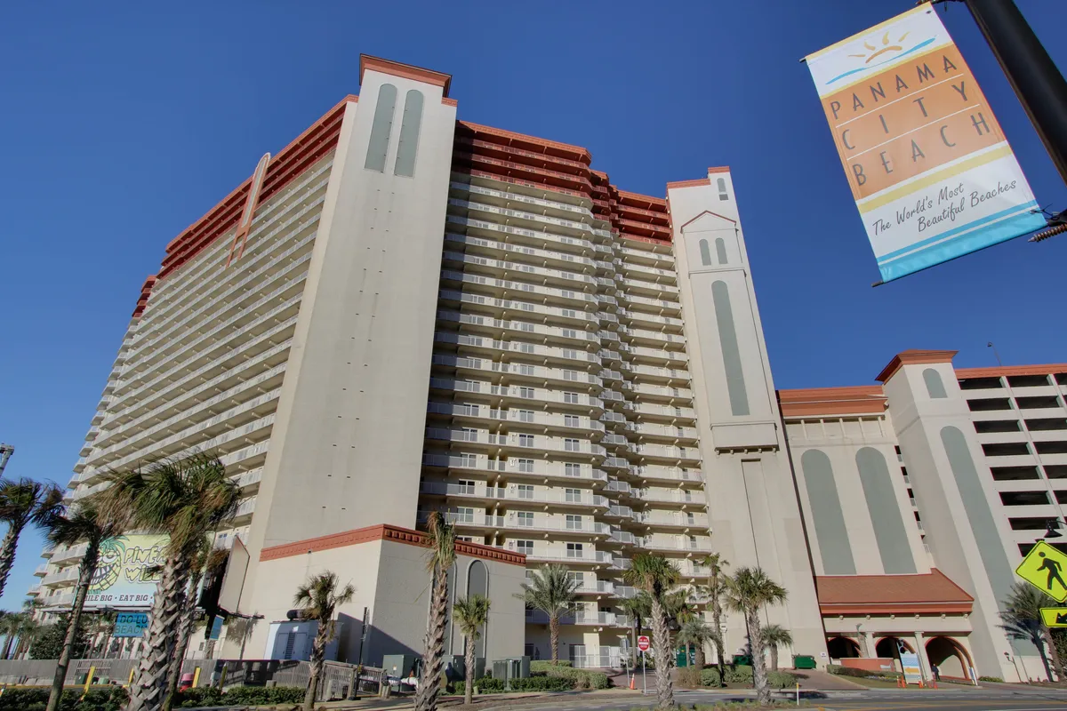 An exterior shot of a multi-story condominium building with inviting balconies, palm trees, and a colorful Panama City Beach banner. The scene emphasizes a resort atmosphere with nearby amenities and easy access to local attractions, suggesting both luxury and proximity to the action.
