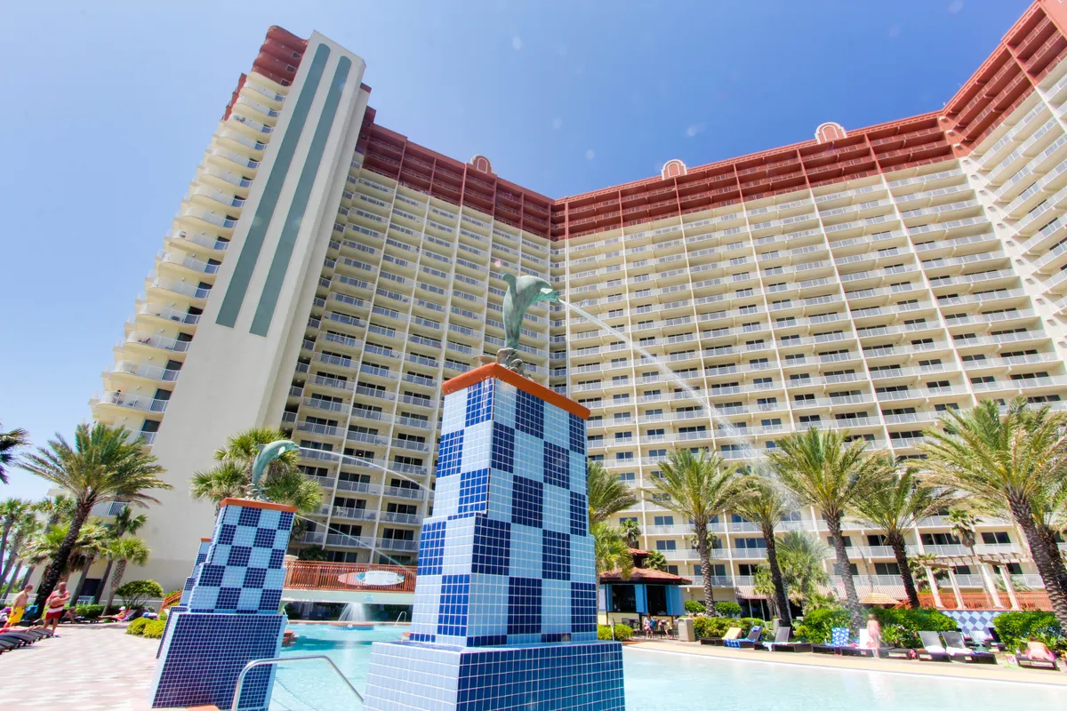 A wide exterior shot of the Shores of Panama resort, featuring the tall multi-story building, blue and white tiled fountains with dolphin sculptures, a spacious swimming pool area lined with lounge chairs, and lush palm trees enhancing the tropical vibe.