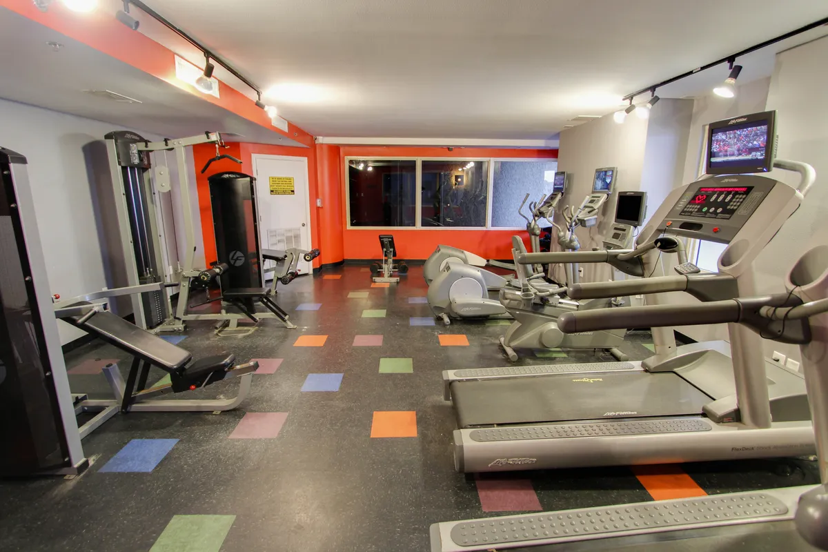 An interior fitness center is equipped with modern treadmills, elliptical machines, and weight training equipment, all set on colorful tiled flooring. Bright lighting and a spacious layout create a motivating environment.