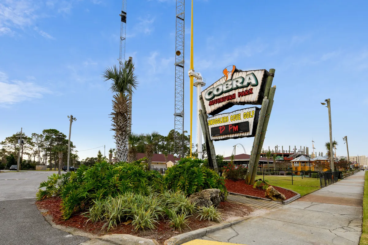 The image features the Cobra Adventure Park sign, a miniature golf course area, tall amusement rides in the background, and lush landscaped greenery with palm trees. The playful atmosphere is unmistakable, with vibrant colors and visible attractions for all ages.