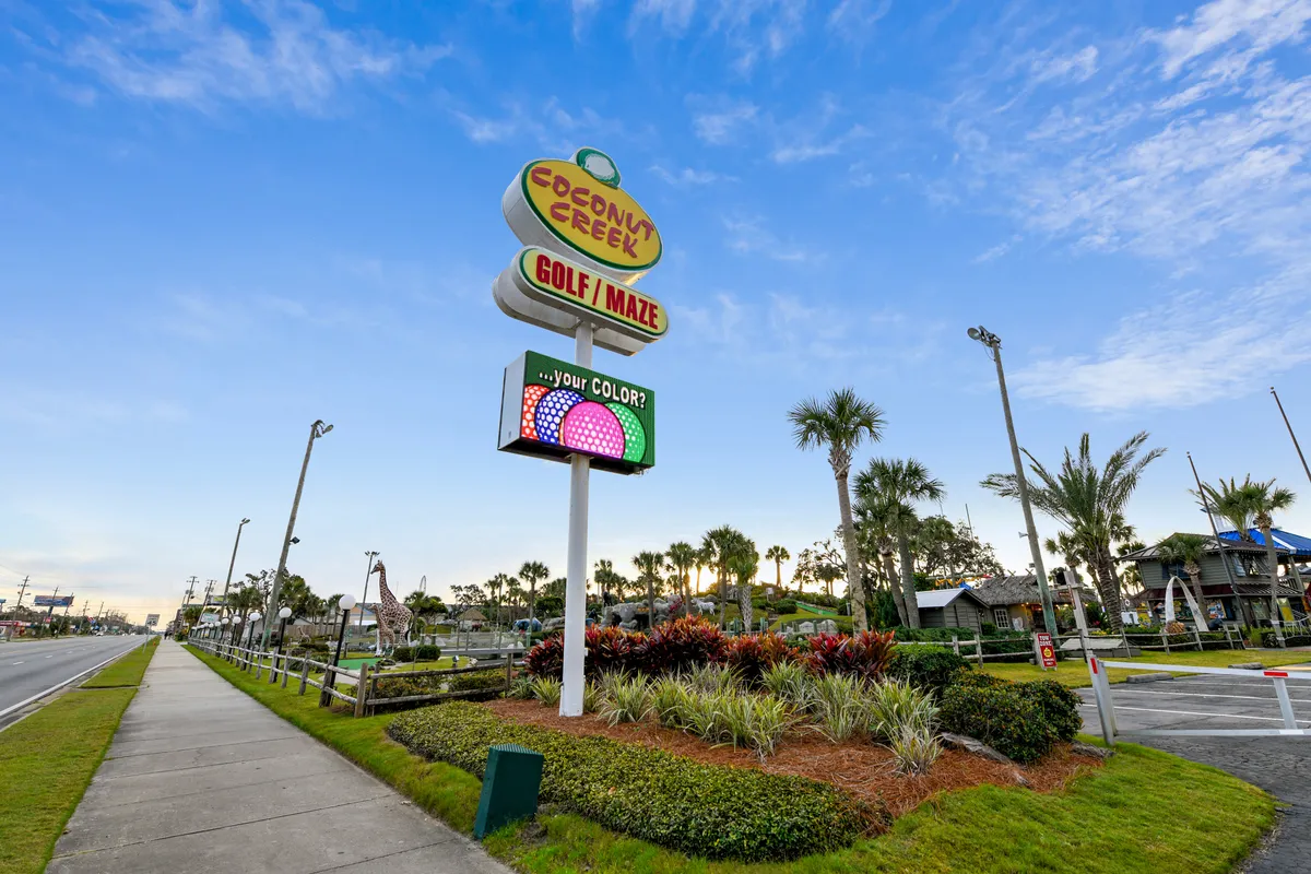 A vibrant miniature golf course is nestled among tall palm trees, with a whimsical giraffe statue and the Coconut Creek Golf / Maze sign visible. Families and groups are seen enjoying playful activities outdoors, surrounded by a fun, engaging atmosphere.