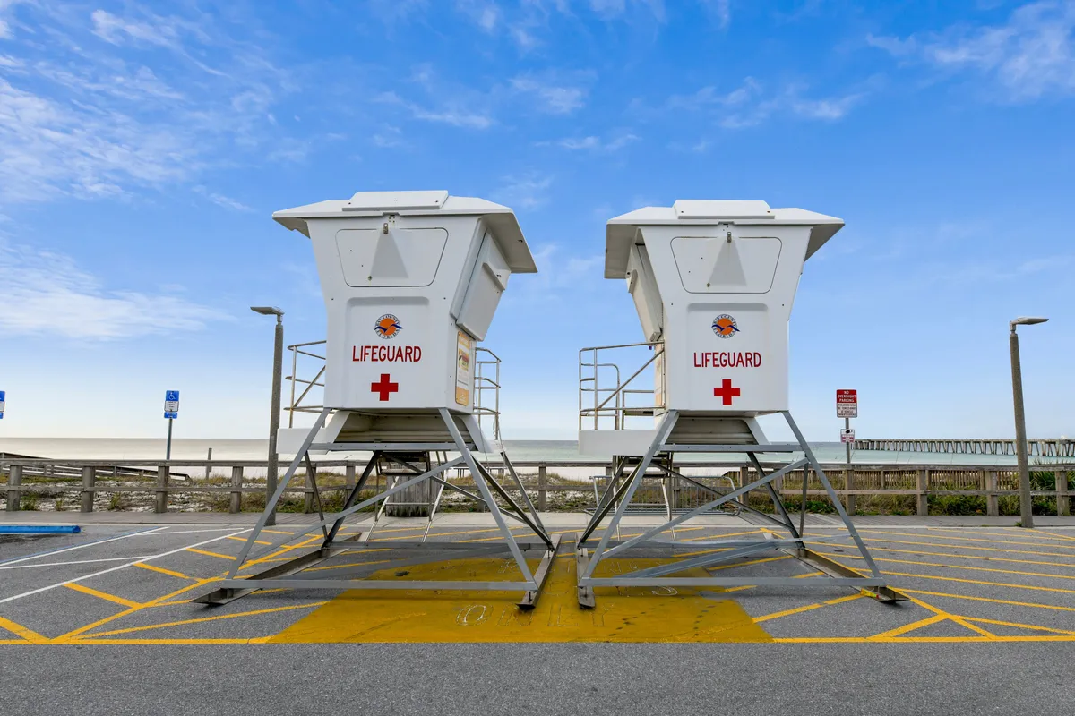 Two white lifeguard towers stand before a wide sandy beach, separated from a parking area by a wooden fence. The sky is bright and blue with scattered clouds, and the scene exudes safety, relaxation, and easy access to the beach.