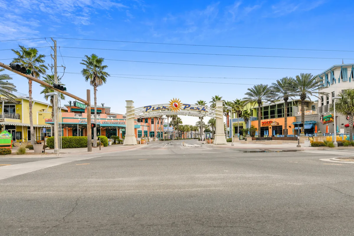This exterior image captures the entrance arch to Pier Park, surrounded by colorful retail and dining buildings, palm trees, and a wide, pedestrian-friendly street bustling with potential visitors. The festive and inviting atmosphere is enhanced by the sun emblem and the vibrant facades, reflecting the area's lively dining and entertainment options.