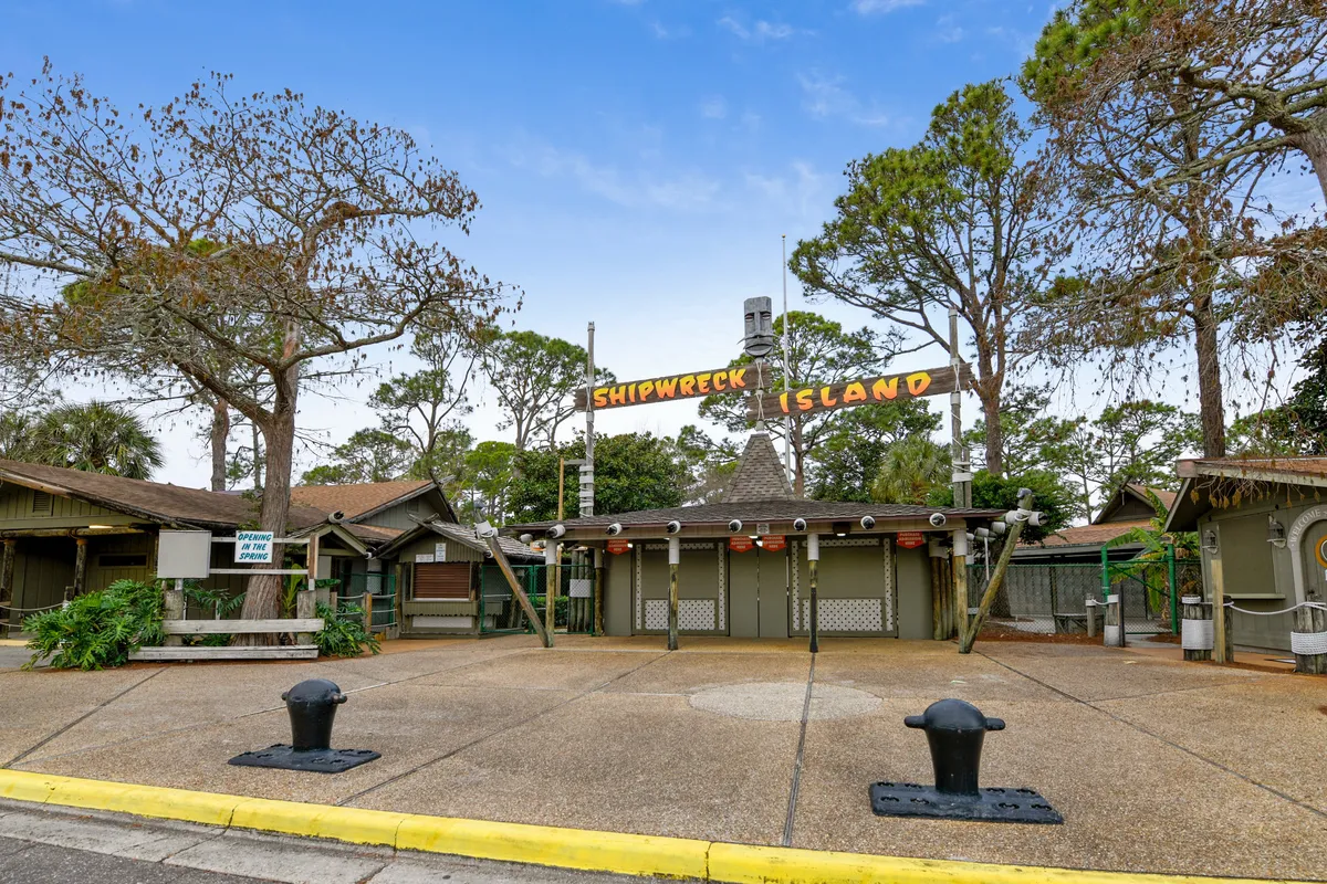 The image displays the entrance sign to Shipwreck Island Water Park, framed by rustic wooden buildings, a large paved open area, and mature trees in the background. The scene captures the anticipation and excitement of a family outing to a signature local attraction.