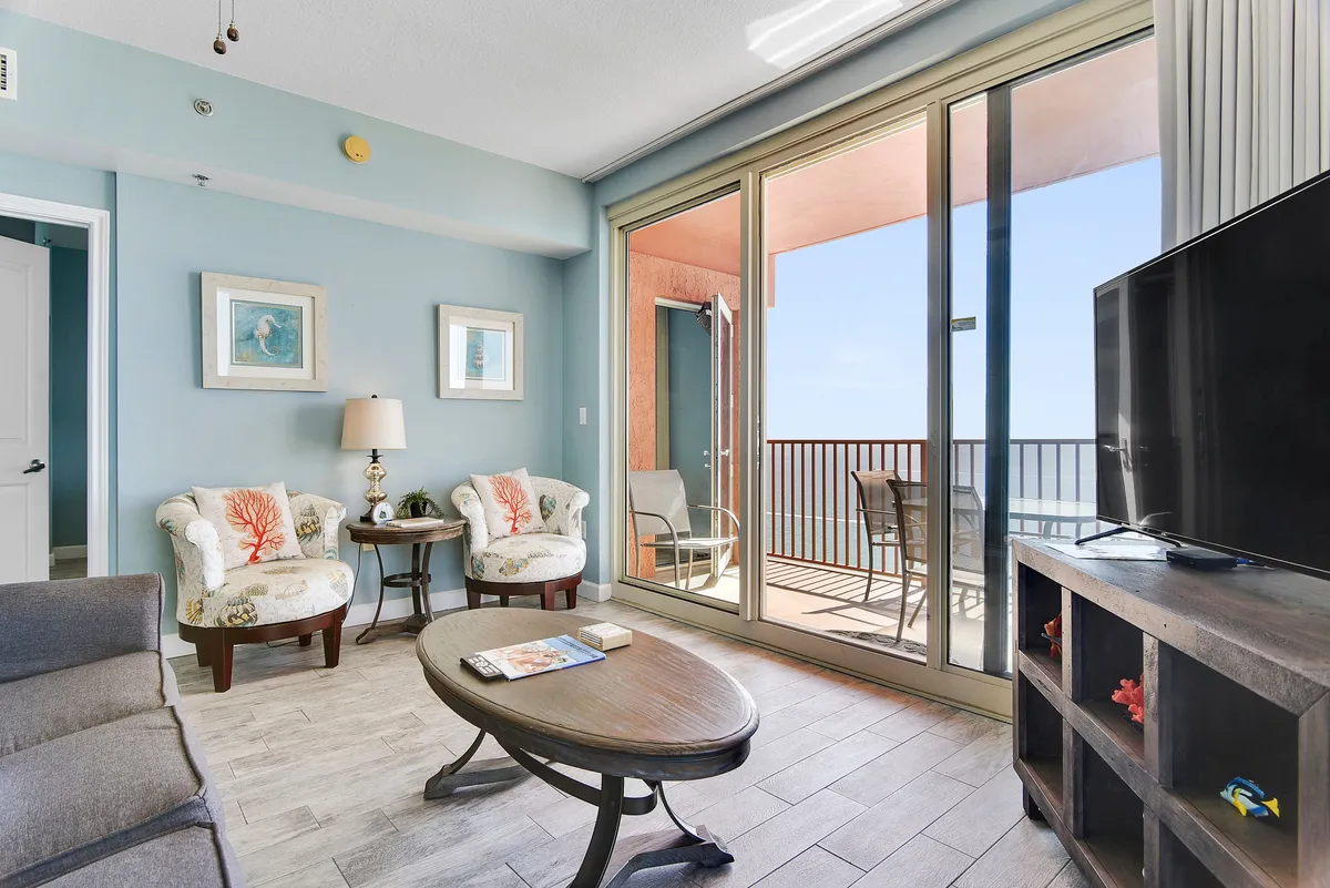 The image shows a living area with large glass doors that open to a wraparound balcony, comfortable seating, and a coffee table with a book. The space is bathed in tranquil morning light, seamlessly blending indoor and outdoor living and offering unobstructed views of the Gulf.