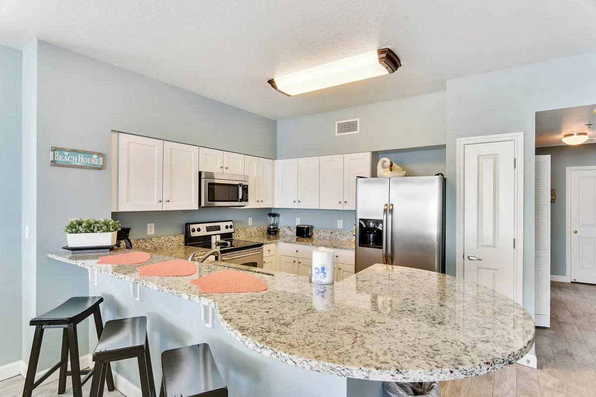 A cheerful kitchen scene centered on a granite-topped island, stainless steel appliances, and a 'beach house' sign that adds personality. The space exudes warmth, with cookware visible and comfortable seating suitable for both cooking and casual gathering.