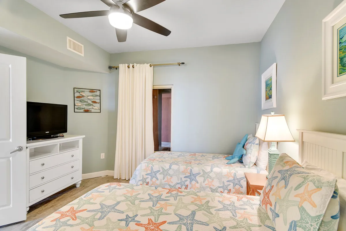 A cheerful bedroom with two twin beds featuring starfish-patterned bedding, a nightstand with a lamp between them, a TV on the dresser, and decorative wall art. The room is bright and inviting, ideal for children or friends traveling together.