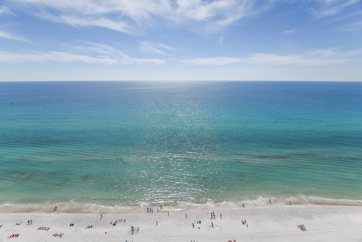 A panoramic view of the sugar-white beach fronting Panama City Beach, with gentle waves lapping at the shore and people enjoying sunbathing, swimming, and beach volleyball. The pristine sand and clear ocean create a quintessential family-friendly beach atmosphere, with easy access directly from the resort.
