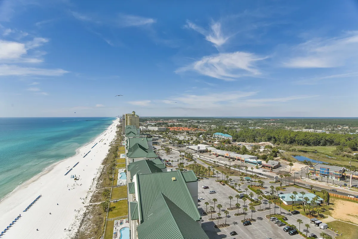 A panoramic view of the turquoise ocean and white sandy beach, with high-rise buildings and palm trees lining the shore. The sky is a clear blue, creating a vibrant and luxurious coastal atmosphere with visible beach and waterfront dining options.