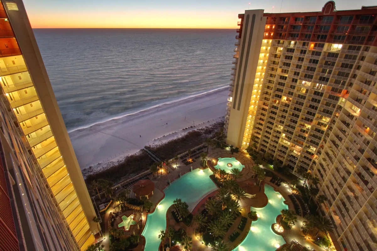 This image captures a stunning sunset over the ocean, with a swimming pool area and beachfront high-rise buildings in the frame. People are seen swimming, walking along the beach, and relaxing by the poolside, all under a serene evening sky.