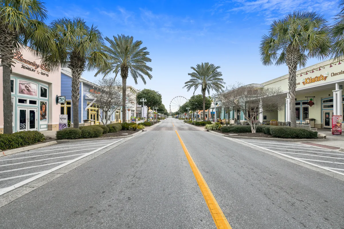 A lively streetscape at Pier Park, featuring palm trees, storefronts, and the Ferris wheel in the distance. People are strolling, shopping, and dining, adding to the energetic atmosphere.