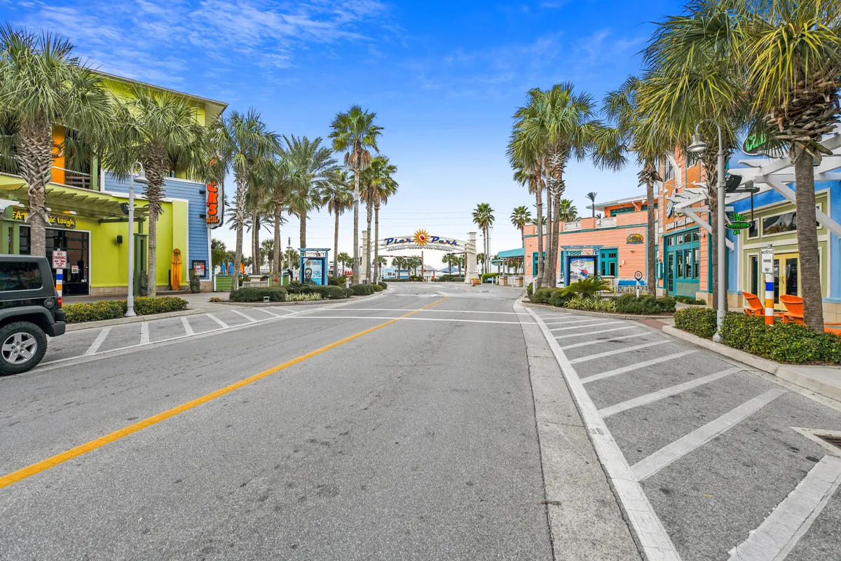A vibrant exterior street scene featuring colorful buildings, palm trees, and an inviting archway sign. People are seen strolling, visiting shops, and enjoying outdoor dining against the backdrop of a glowing sunset.